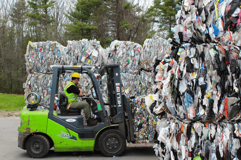 A man surrounded by various bails operates a green colored forklift in a recycling bail yard with trees in the backyard